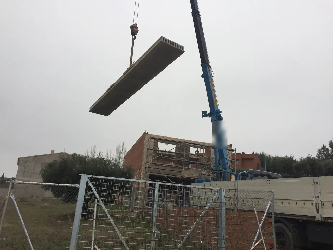 Izado de una placa alveolar con grúa móvil sobre la obra, con el camión de transporte a la derecha
